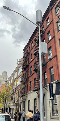 A streetlight pole in NYC has a box and cylinder shaped device attached to the top of the pole.